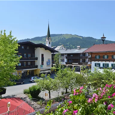 A picturesque landscape with traditional buildings and a tall church tower. In the background, the mountains stretch out under a clear blue sky.