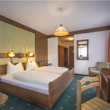 A cozy bedroom with a double bed, two pillows, and wooden details. There is a table, chairs, and a window with curtains.