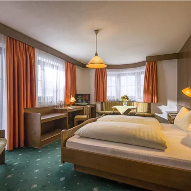 A cozy hotel room with two beds and a desk. Large windows with curtains provide plenty of daylight.