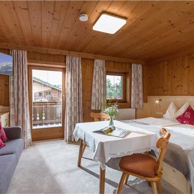 A cozy room with a wooden ceiling and two beds. There is a small table, a television, and windows looking outside.