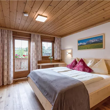 A cozy bedroom with a wooden ceiling and warm colors. The room has a large window and decorative cushions on the bed.