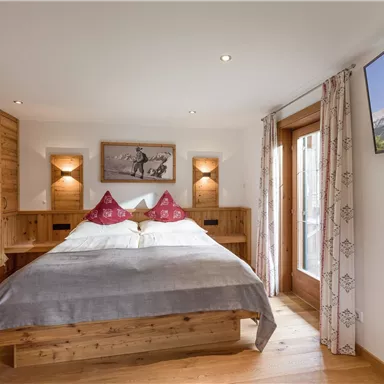 A cozy bedroom with a wooden bed and decorative cushions. Large windows let in a lot of light and offer a beautiful view of the surroundings.