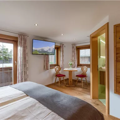 A cozy bedroom with a large bed and a television. There is a small dining area with two chairs and windows that let in a lot of light.