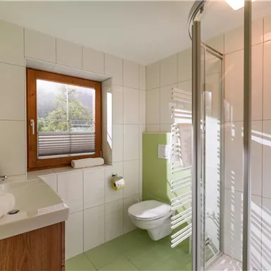 A modern bathroom with a shower and a window. The walls are light tiled, and there are colorful accents in green.
