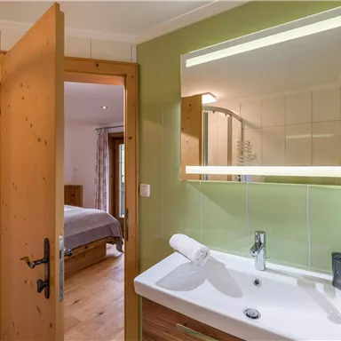A modern bathroom with a sink and large mirror. In the background, there is a specified access to a bedroom.