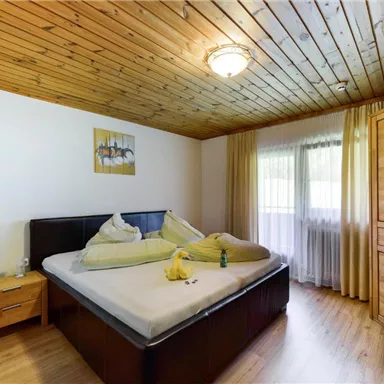 A cozy bedroom with a large bed and a wooden ceiling. Natural light flows through the windows, which are framed by light curtains.