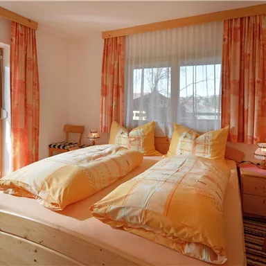 Ein helles Zimmer mit zwei Betten, großen Fenstern und warmen Farbtönen. Die Möbel sind aus Holz und es gibt einen kleinen Tisch mit Stühlen.