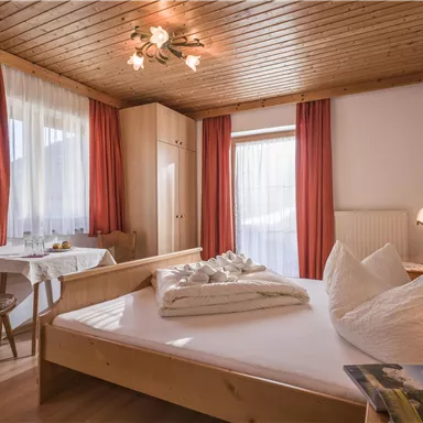 A cozy bedroom with a double bed and gentle wooden furniture. Large windows provide plenty of daylight and a pleasant atmosphere.