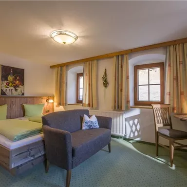 A cozy hotel room with a double bed, an armchair, and a small desk. The walls are painted in warm colors, and large windows let in plenty of daylight.