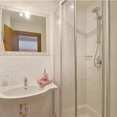 Ein modernes Badezimmer mit einer Dusche und einem Waschbecken. Die Wände sind hell gefliest, und es liegen rosa Handtücher auf dem Waschbecken.