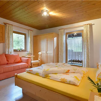 A cozy bedroom with a double bed, a sofa, and wooden shelves.  
Large windows let in plenty of light, and the decor is friendly and inviting.
