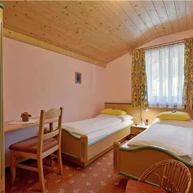 A cozy room with two single beds and a small table. The walls are pink and the windows have colorful curtains.