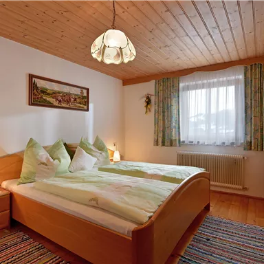 Ein gemütliches Schlafzimmer mit einem Doppelbett und angenehm hellem Holzinterieur. Große Fenster sorgen für viel Tageslicht und eine einladende Atmosphäre.