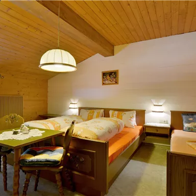 A cozy bedroom with wooden walls and a sloped roof. There are two beds, a table, and pleasant lighting.