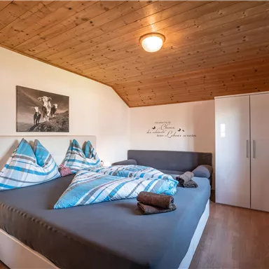A cozy bedroom with a double bed and light bedspreads. The walls are in soft colors and there is a window with red curtains.
