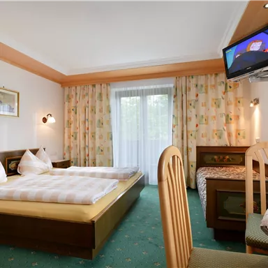 Ein gemütliches Hotelzimmer mit einem Doppelbett und einem Fernseher. Das Zimmer hat große Fenster und eine freundliche Farbgestaltung.
