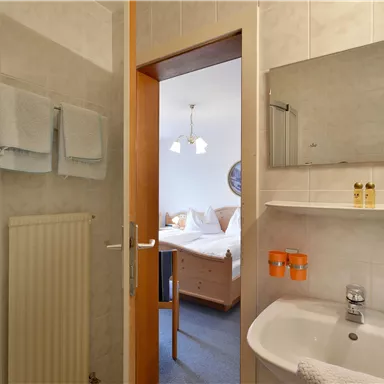 Ein Badezimmer mit Dusche, Waschbecken und Spiegel. Im Hintergrund ist ein Schlafzimmer mit einem Bett sichtbar.