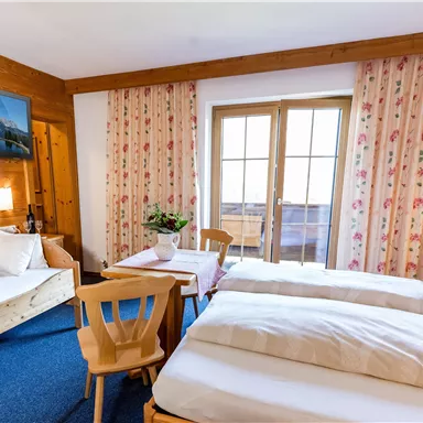 Ein gemütliches Hotelzimmer mit zwei Einzelbetten und einem Schlafsofa. Große Fenster mit Blick auf einen Balkon und ein schönes Holzdekor prägen die Atmosphäre.