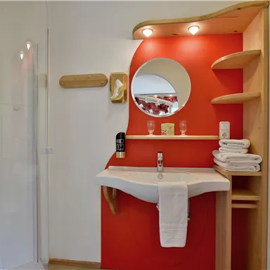 Ein modernes Badezimmer mit einer Dusche und einem Waschbecken. Die Wände sind teilweise rot gestrichen und es gibt Holzregale.