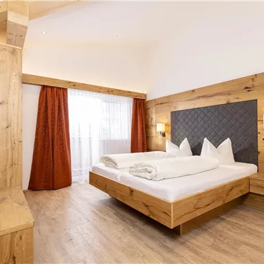 A modern bedroom with wooden paneling and a large bed. The windows let in plenty of daylight, and the curtains are in warm orange tones.