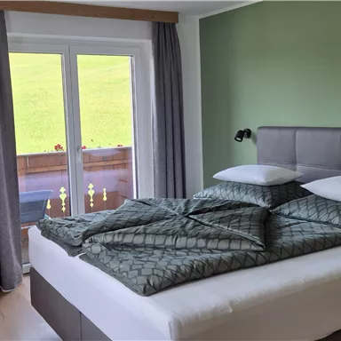 Ein modernes Schlafzimmer mit einem großen Bett und grünen Wänden. Große Fenster lassen viel Tageslicht herein und bieten einen Blick ins Freie.