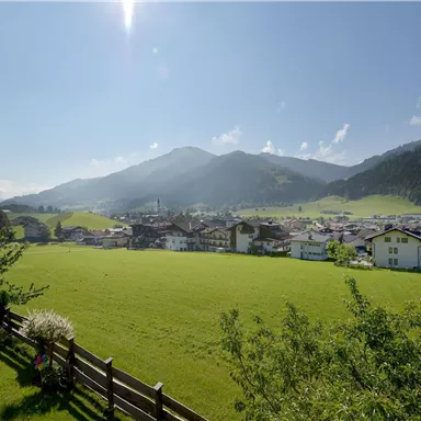 A picturesque landscape with green meadows and a small village. In the background, tall mountains and a clear blue sky can be seen.