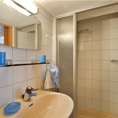 Ein modernes Badezimmer mit einem Waschbecken, einem Spiegel und einer Dusche. Die Wände sind gefliest und es gibt einige Badezubehörteile auf dem Regal.