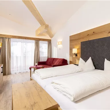 A modern hotel room with wooden interiors and a cozy double bed. The room features a seating area, a television, and large windows with daylight.