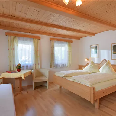 Ein gemütliches Schlafzimmer mit Holzdecke und zwei Betten. Der Raum ist hell und freundlich mit großen Fenstern und einem kleinen Tisch.