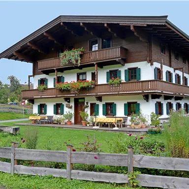 Ein traditionelles Chalet im alpenländischen Stil mit Balkonen und Blumenkästen. Darum herum befindet sich eine grüne Wiese und ein Holzzaun.