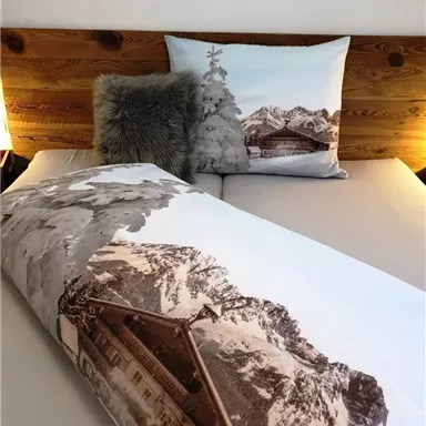 A cozy bed with a duvet and pillows in mountain design. In the background, wooden elements and soft light can be seen.