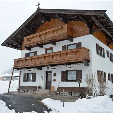 A traditional Alpine house with balconies, surrounded by snowy terrain. The sky is cloudy, and light snow is falling.
