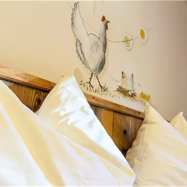 A cozy bedroom with two large white pillows and a chicken painting on the wall. The decor creates a relaxed, rural atmosphere.