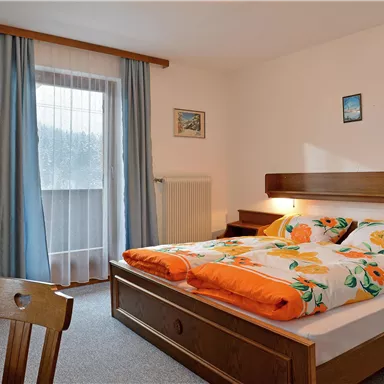A cozy bedroom with a double bed and colorful quilts. The room has large windows with curtains and a simple wooden desk.