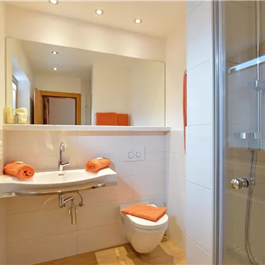 A modern bathroom with bright tiles and a large mirror. Orange towels are placed on the sink, and there is a comfortable shower.