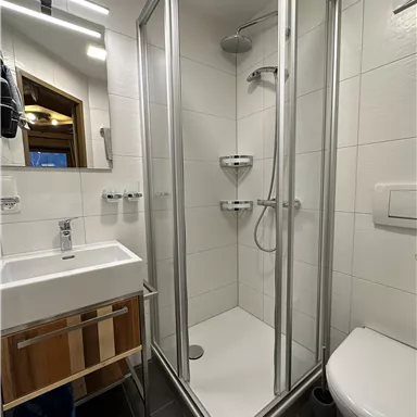 Ein modernes Badezimmer mit einer Dusche, einem Waschbecken und einer Toilette. Die Wände sind hell gefliest und es gibt eine große Spiegel.