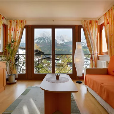 A cozy living room with an orange sofa and a small table. Large windows provide a view of the mountains and let in plenty of light.