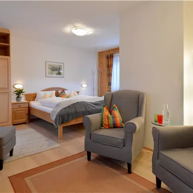 A cozy bedroom with a double bed, two comfortable armchairs, and a television. The room is bright and friendly furnished, with a carpet and a beautiful view through the window.
