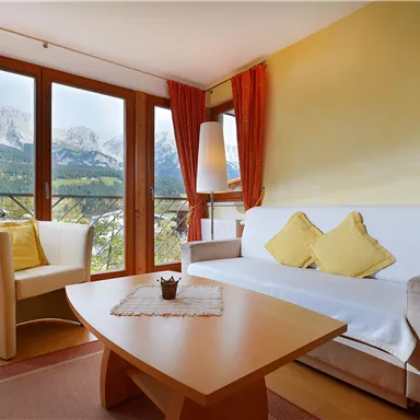 A cozy living room with a white sofa and two yellow cushions. Through the windows, you can see the mountains in the background.