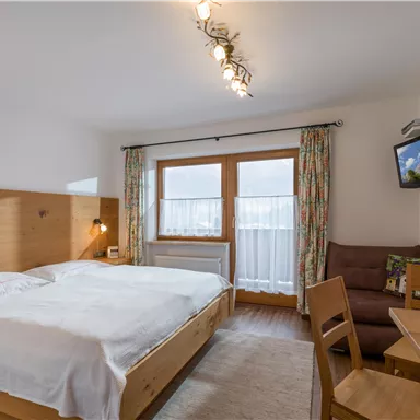 A modern bedroom with a double bed and wooden furniture. There is a window with curtains and a small desk with a chair.