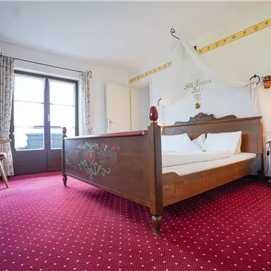 A cozy bedroom with a large bed and traditional furniture. The room has a red carpet and windows overlooking the outside.