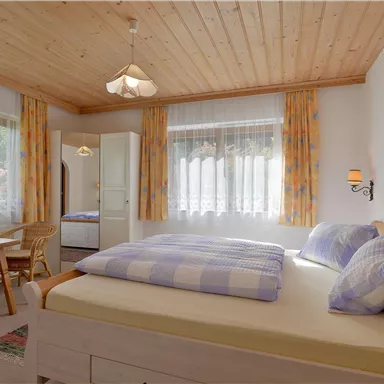 A cozy bedroom with wooden ceilings and large windows. The room features a double bed and a table with chairs.
