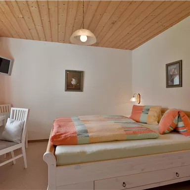 A cozy bedroom with a double bed and colorful blankets. The room has a desk and windows with curtains.