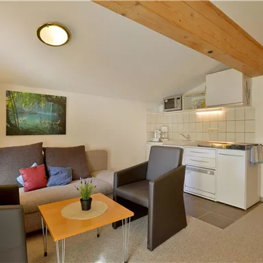 A cozy living kitchen with a sofa, two chairs, and a small table. The decor is modern and bright, with a painting on the wall.