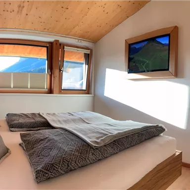Ein modernes Schlafzimmer mit einem großen Bett und gemütlicher Bettwäsche. An der Wand hängen ein Fernseher und ein Spiegel, und durch die Fenster scheint das Licht.