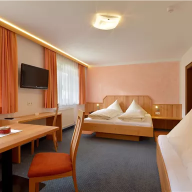 A modern hotel room with a double bed and a seating area. There is a table, chairs, and a television, surrounded by large windows.
