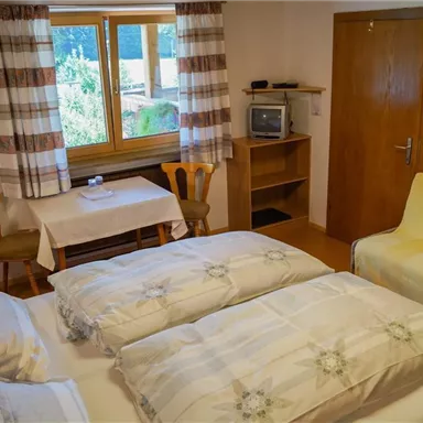 A cozy room with a double bed, a table, and chairs. The room has windows overlooking greenery and a comfortable atmosphere.