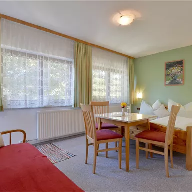 Ein gemütliches Zimmer mit zwei Betten, einem Tisch und Stühlen. Große Fenster sorgen für viel Tageslicht und eine angenehme Atmosphäre.