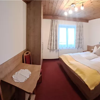 A cozy room with two beds, wooden ceiling, and carpeted floor. A small table and a window with curtains complete the furnishings.