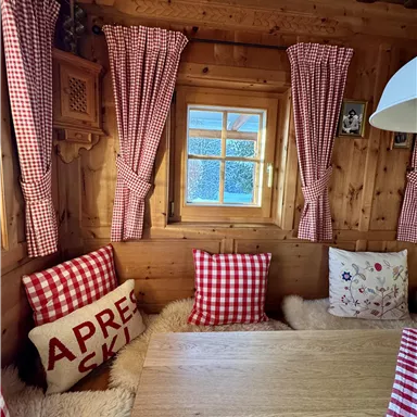 Eine gemütliche Holzstube mit karierten Vorhängen und Kissen. Am Tisch steht ein Fenster mit Blick nach draußen.